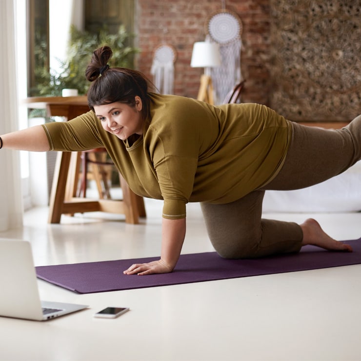 Female of size practicing yoga indoors on mat by watching online video lesson on laptop computer on floor