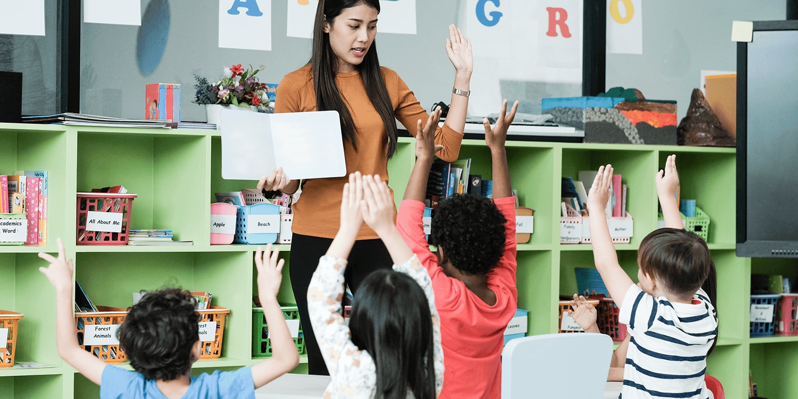 Several students raising their hands to a book a teacher is holding up