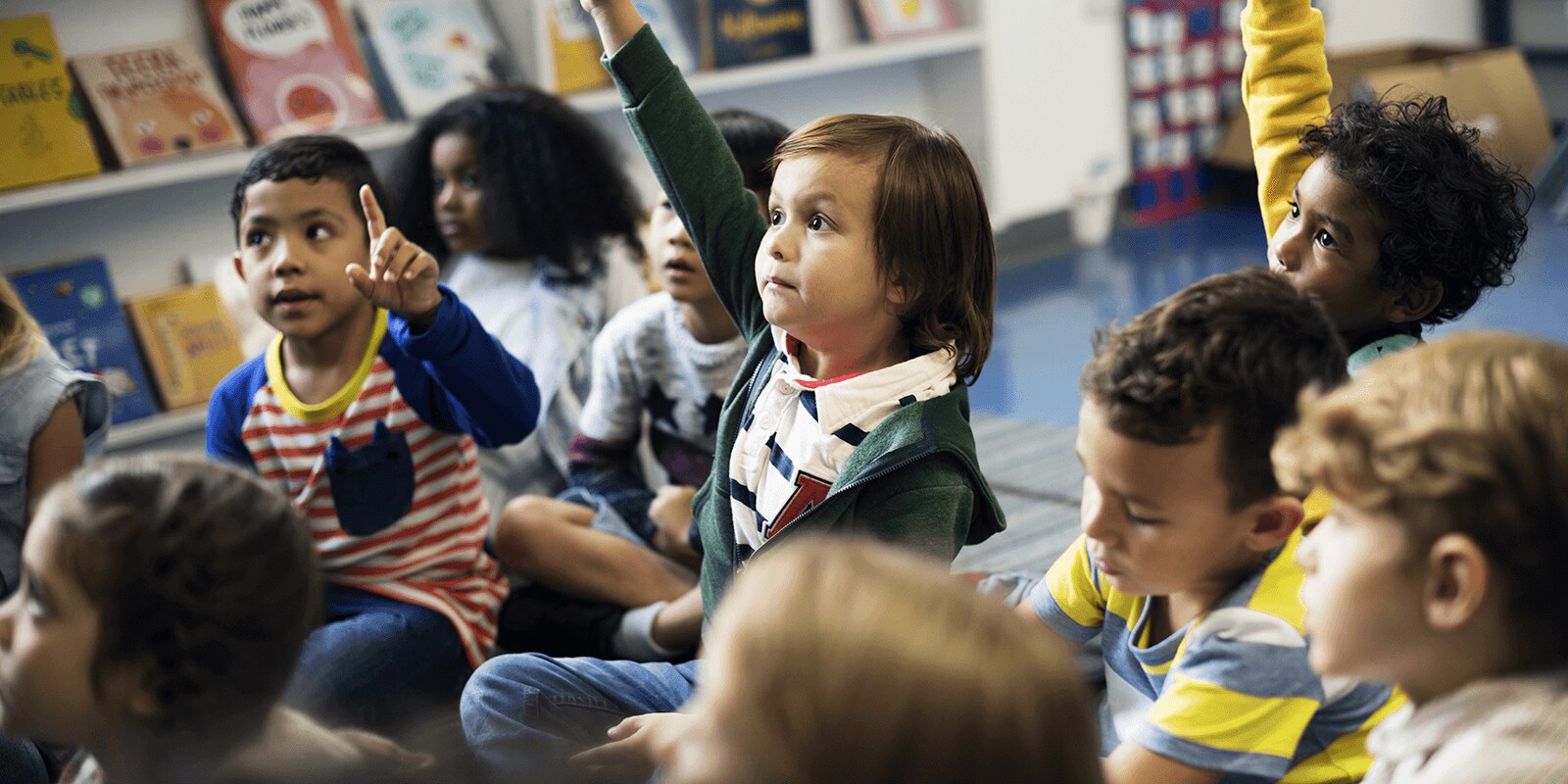 Grade school children in a classroom raising their hand. 