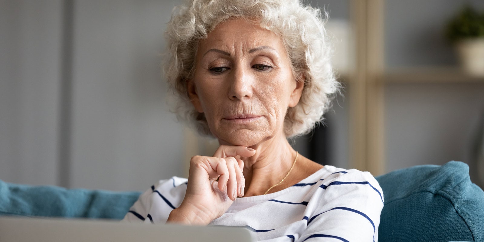 An elderly woman with curly white hair looks intently at laptop while resting her chin on her hand.
