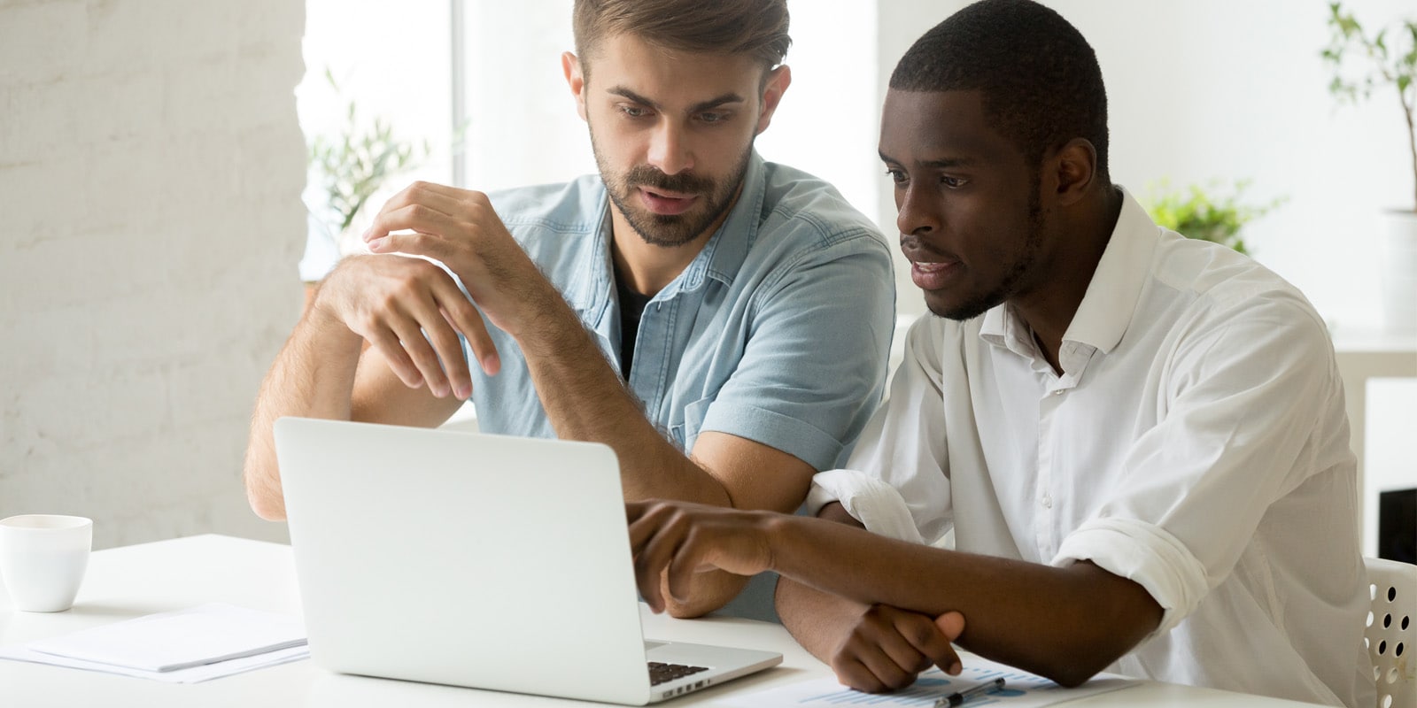 Two men, one in a white shirt and the other in a blue shirt, sit at a table working together on a laptop in a bright office.