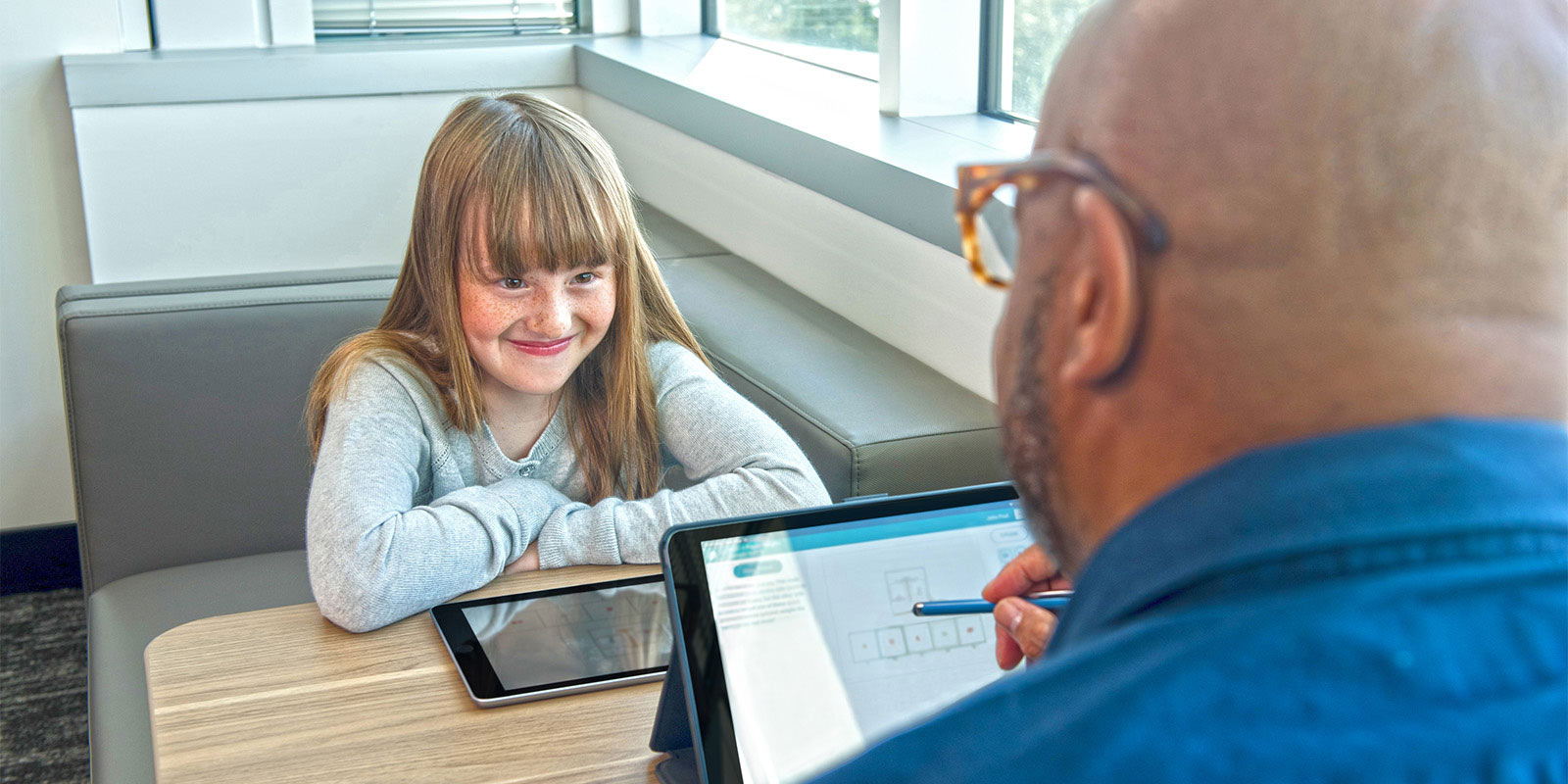 During an assessment, a young girl smiles at a man across the table; both have tablets.