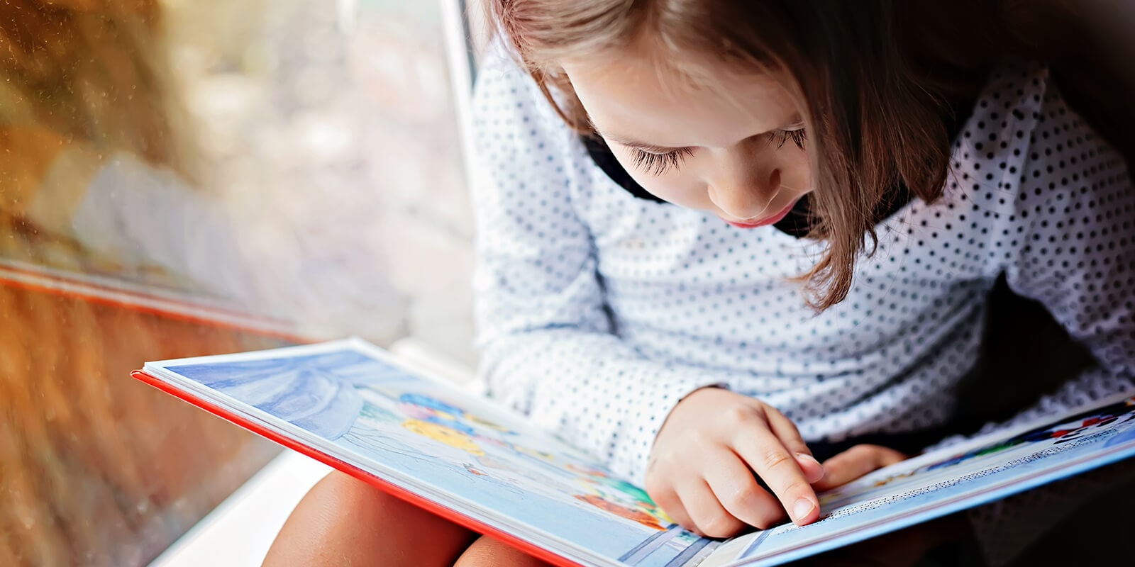 Young child in a polka-dot shirt reading a colorful book by a window.