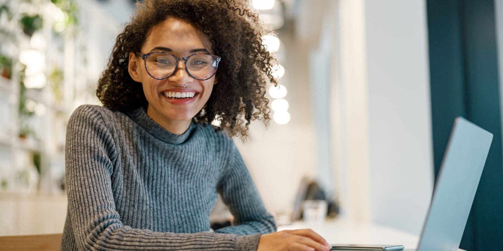 Person in a gray sweater working on a laptop in a bright, plant-decorated cafe setting.