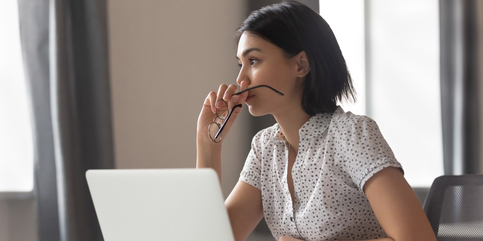 A woman in a polka dot blouse sits at a table in a room, using a laptop and holding a pair of eye glasses.