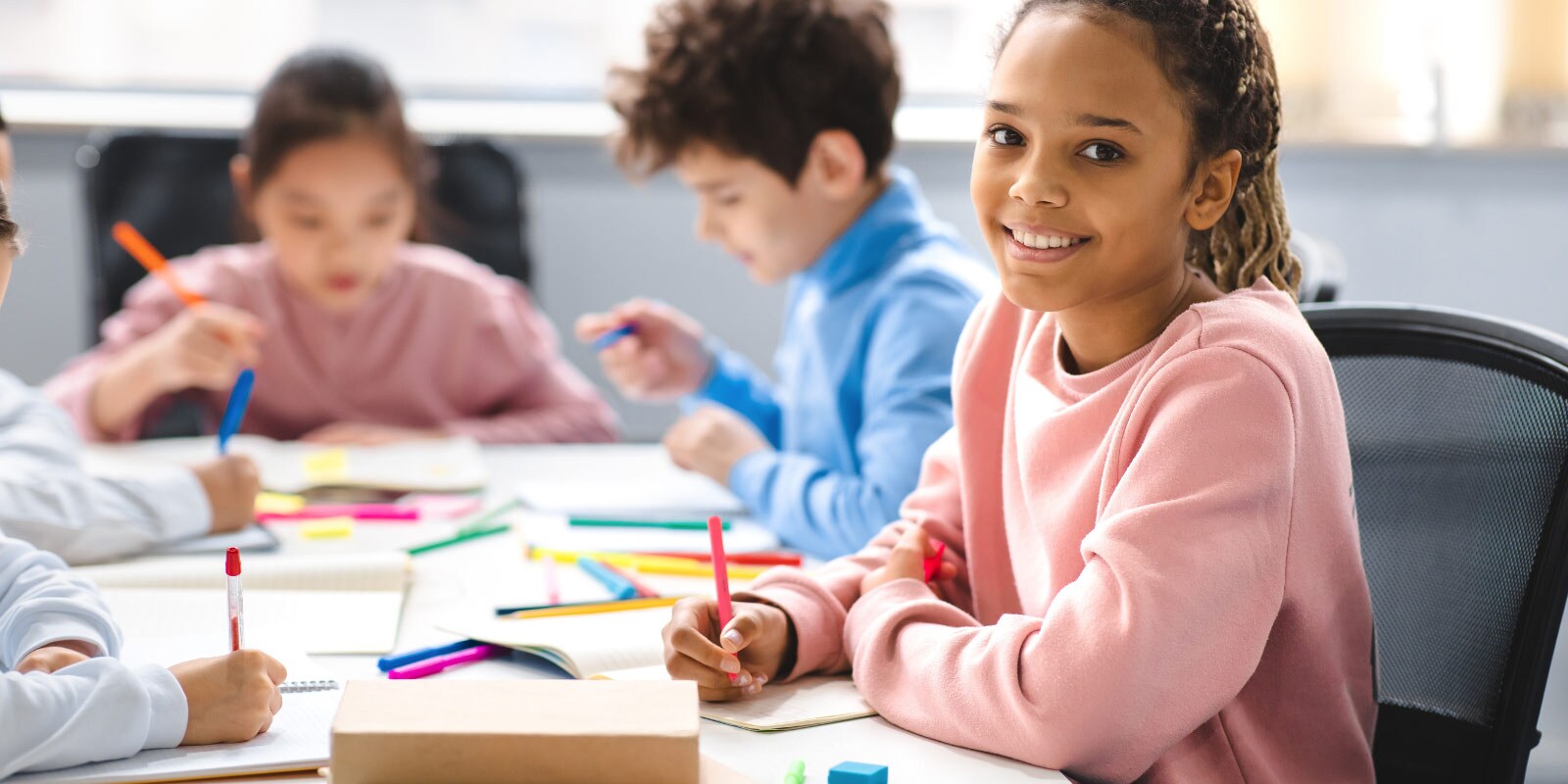 Children seated at a table in a classroom, working on colorful papers and drawings with pencils and crayons scattered around.