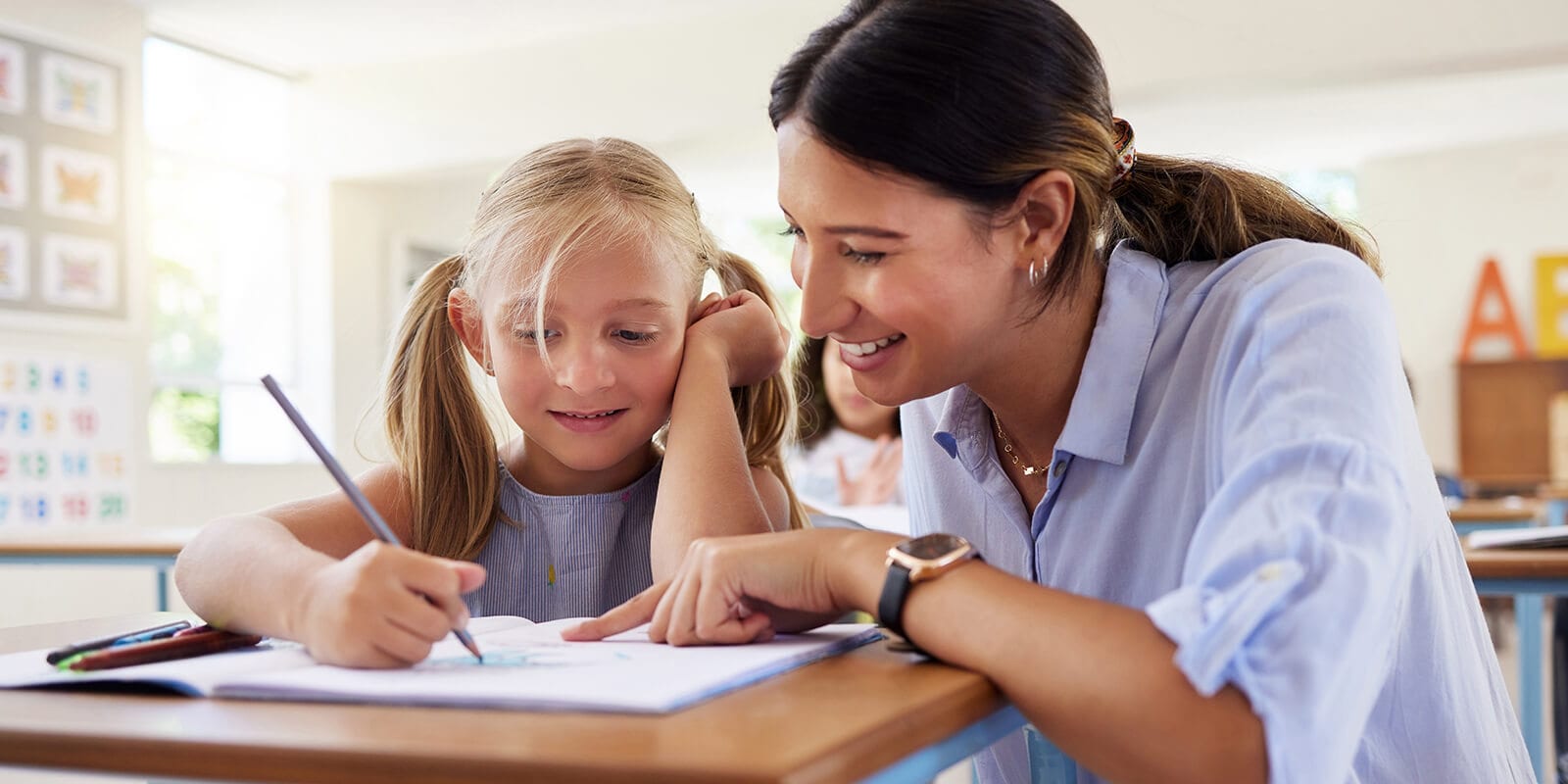 A teacher leans close to assist a young student with her work in a bright, colorful classroom setting.