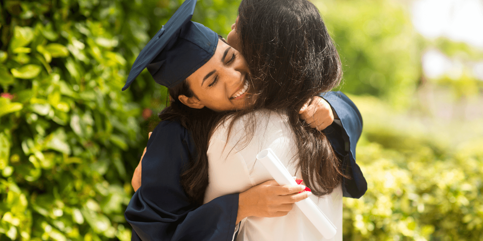 A female graduate in a blue cap and gown hugs another woman outdoors, surrounded by lush greenery, expressing joy.