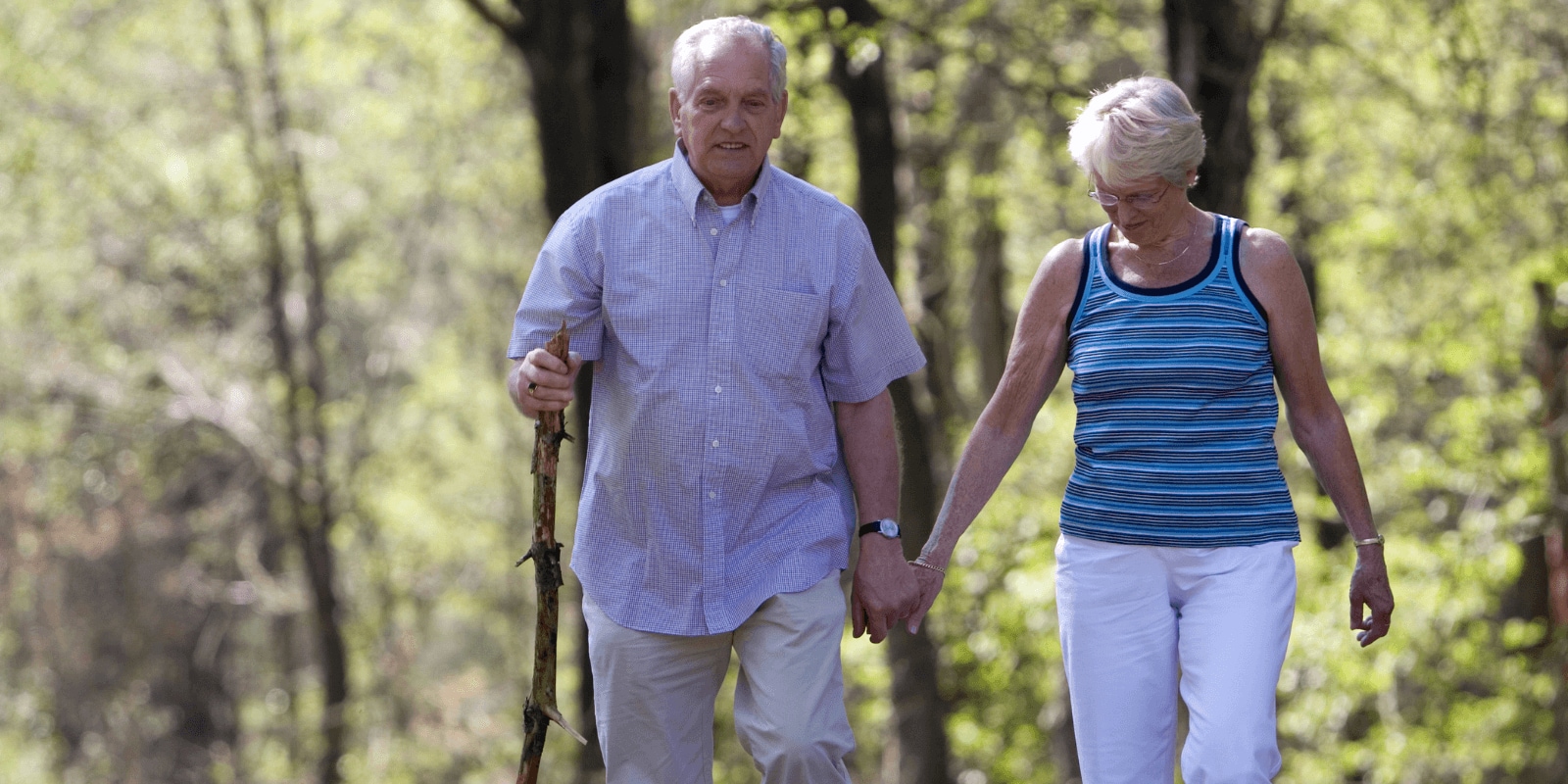 Elderly couple walking in a park, man with cane, woman in tank top, both focused ahead, surrounded by trees in daylight.