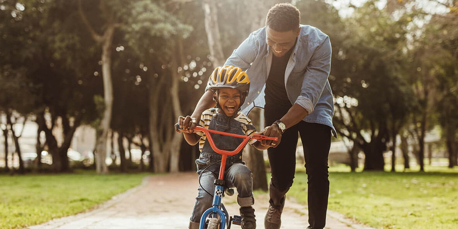 A young child wearing a helmet rides a small blue bike, assisted by an adult in a park on a sunny day.