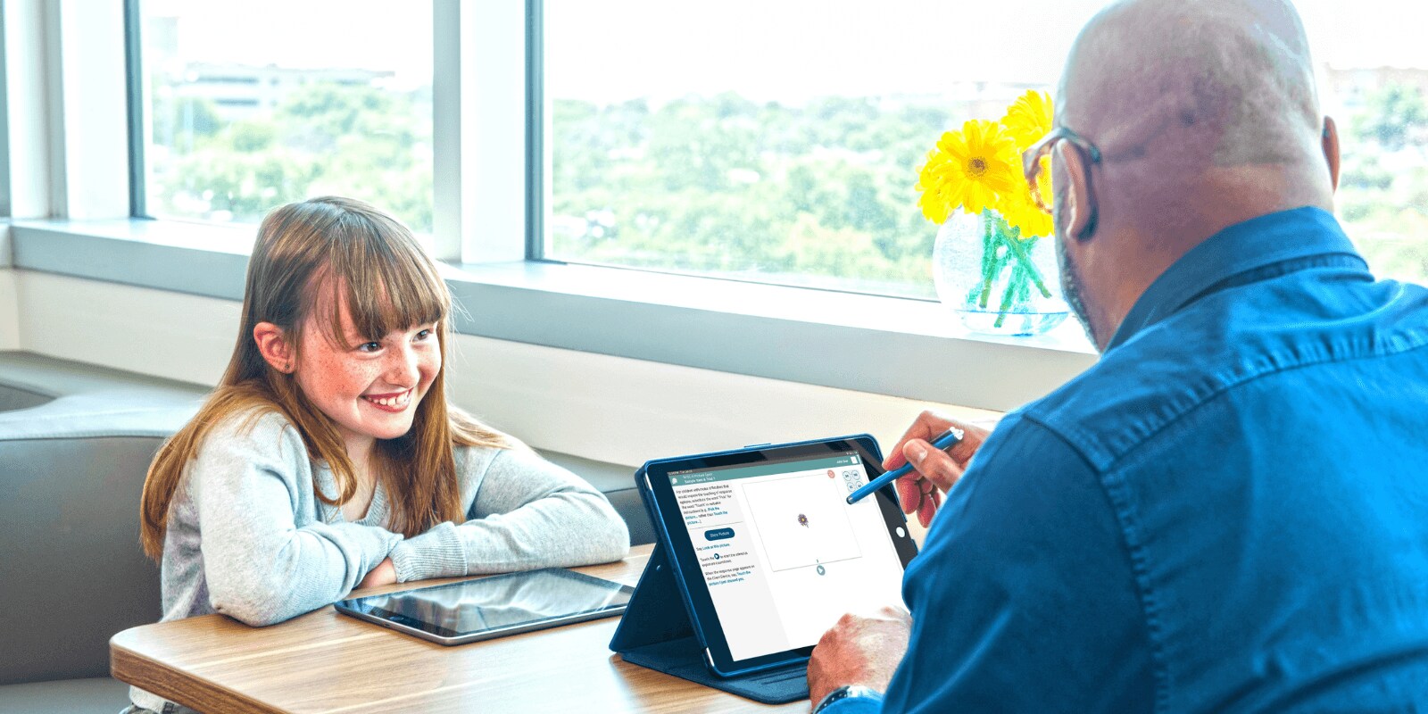 A man and a young girl sit at a table by a window, using digital tablets, with a vase of yellow flowers between them.