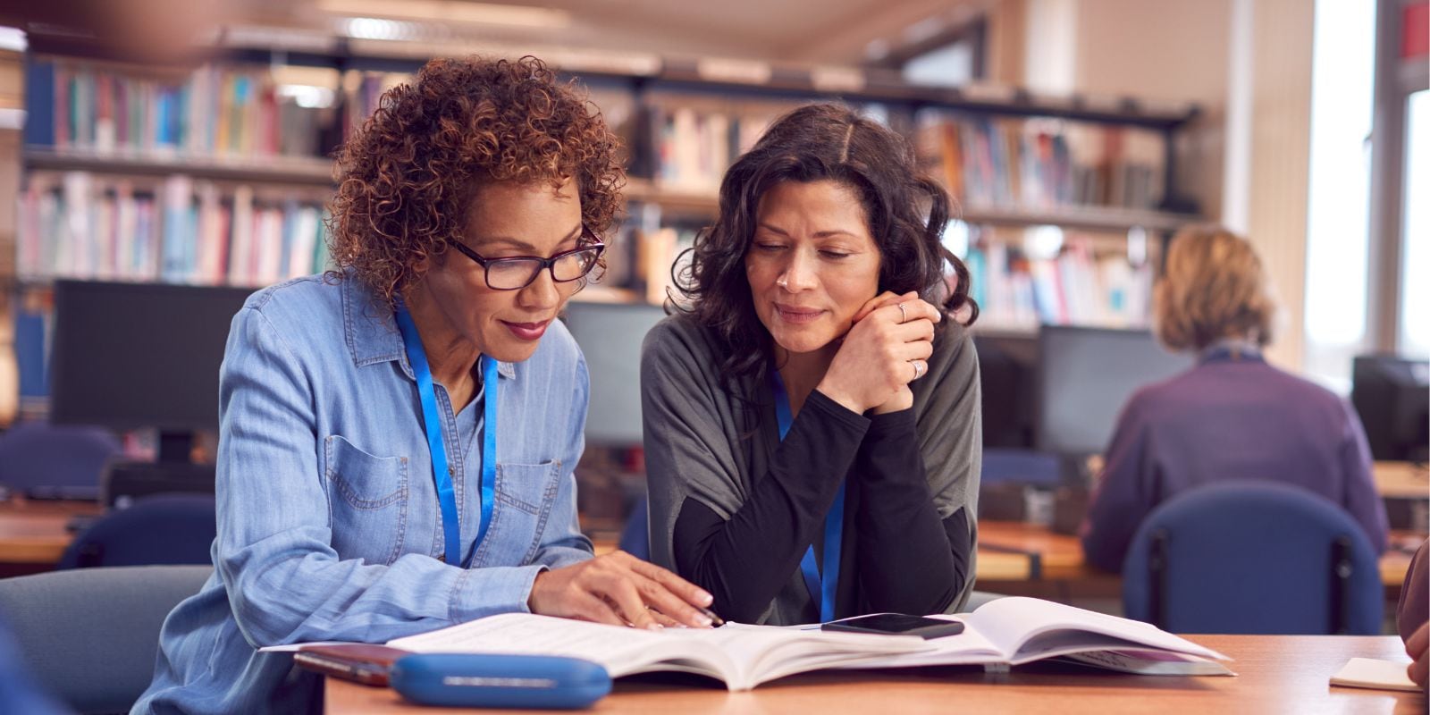 Two individuals are seated at a table in a library, studying a large open book together, with shelves of books and other patrons in the background.