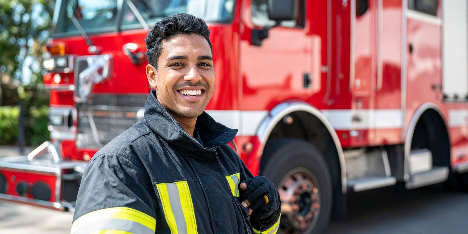 Firefighter in uniform standing in front of a red fire truck, holding a walkie-talkie.