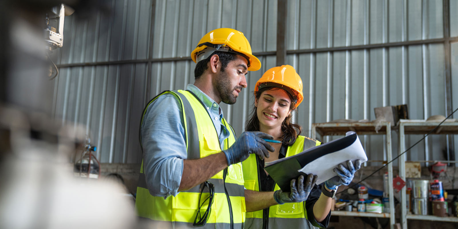 Two workers, male and female, in reflective vests and hard hats discuss over a clipboard in an industrial setting.