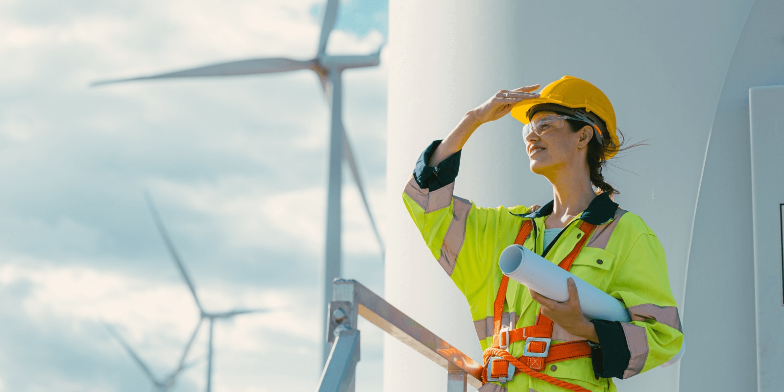 A worker in a high-visibility vest and hard hat stands near a wind turbine, holding blueprints and shielding eyes from the sun.