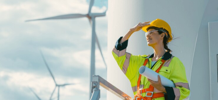 A worker in a high-visibility vest and helmet holds blueprints near a large wind turbine under a clear sky.