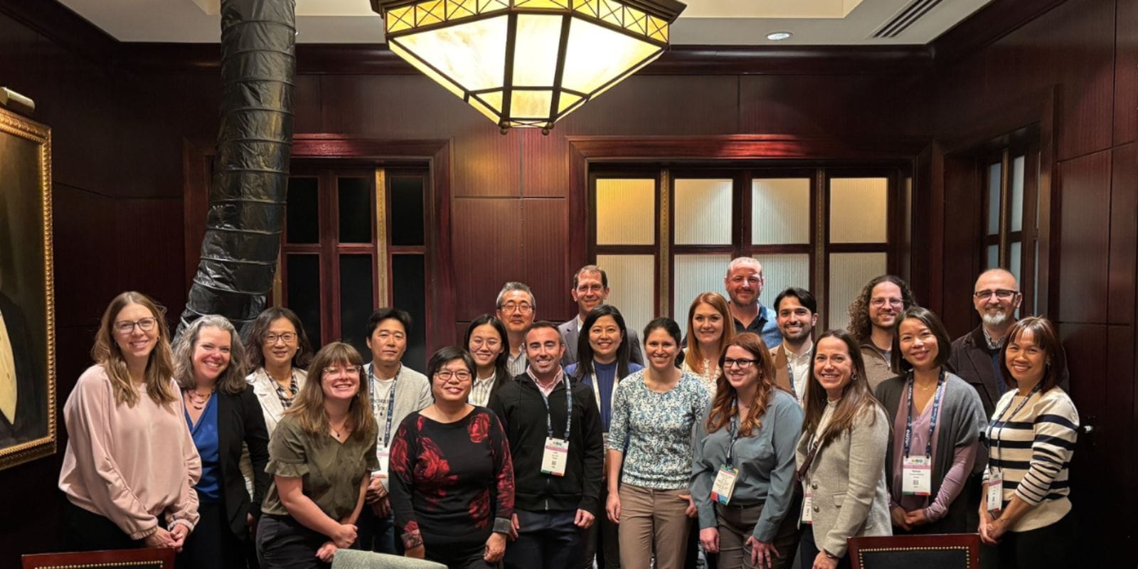 Group of professionals smiling for a photo in a conference room with elegant wood panels and a decorative ceiling lamp.