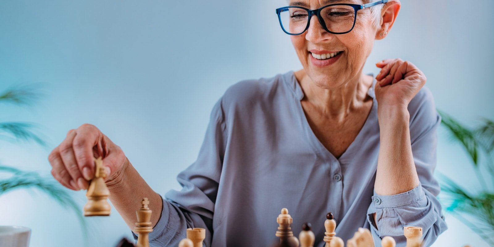 An older woman in a gray blouse is seated at a table playing chess.