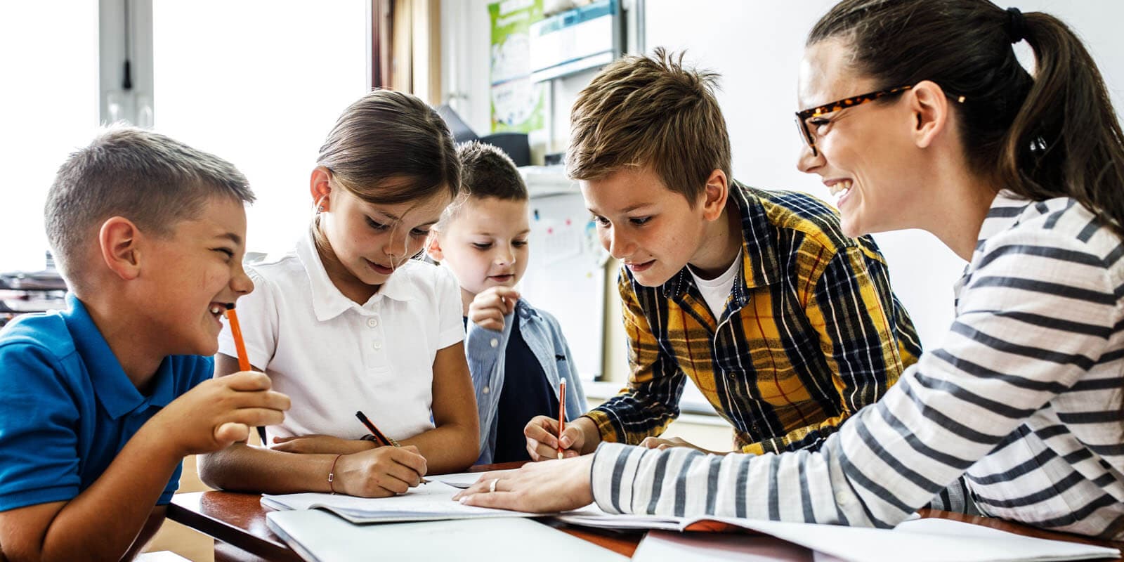 Four students sit at a desk in a classroom, collaborating on a project with notebooks and pens, in a brightly-lit room.
