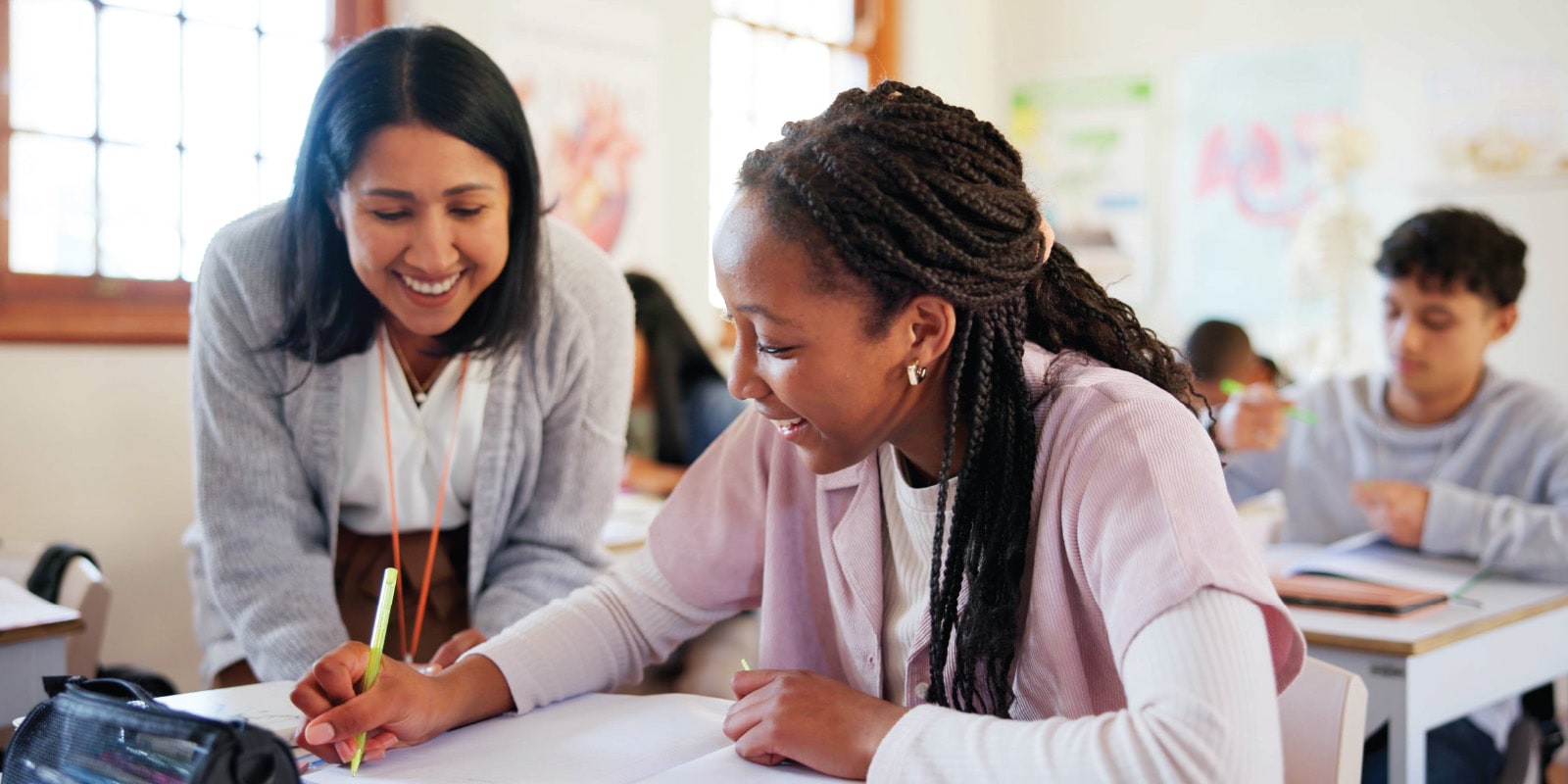 Students in a classroom setting, working on assignments at desks, teacher is assisting a student on assignment.