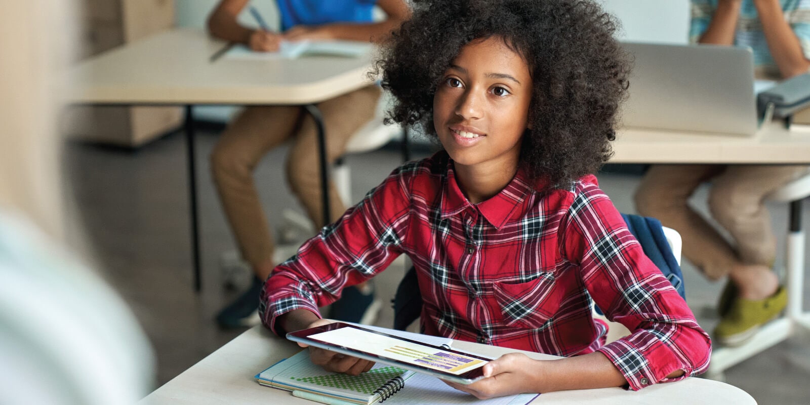 A classroom scene with a child in a red plaid shirt using a tablet at a desk, surrounded by other students working on laptops.