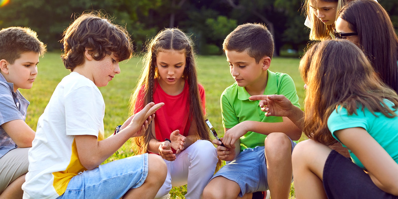 Kids learn about environment during field trip. Students have interesting biology class in nature. School children together with teacher sit down on green grass, search for insects, look through loupe